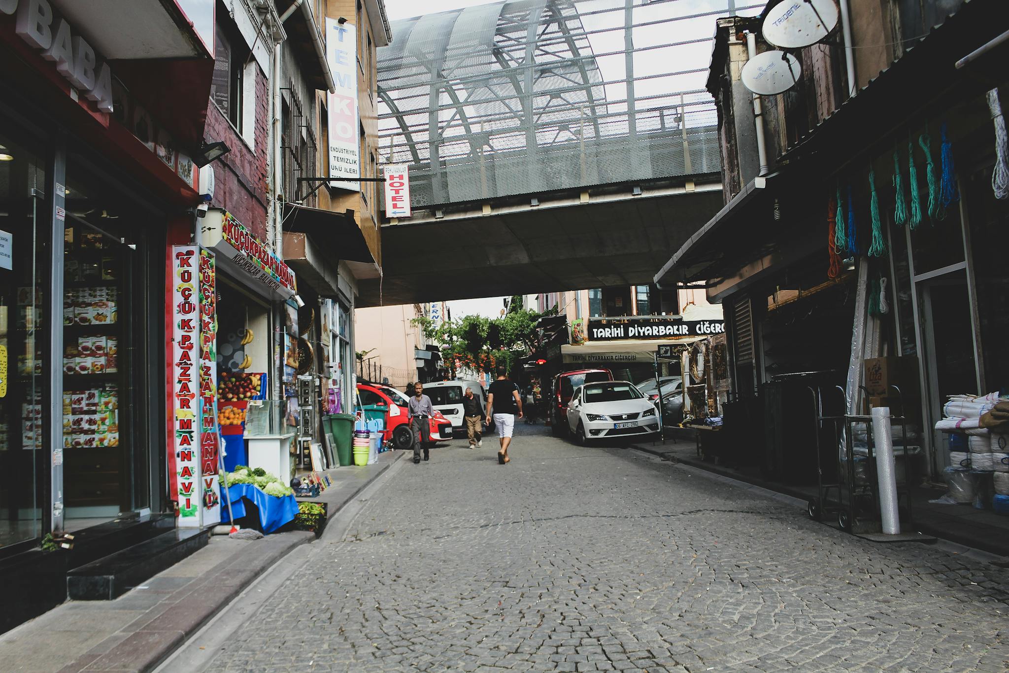 Urban street scene in Kocasinan, Kayseri, Turkey, with shops and pedestrians on a cobblestone road.